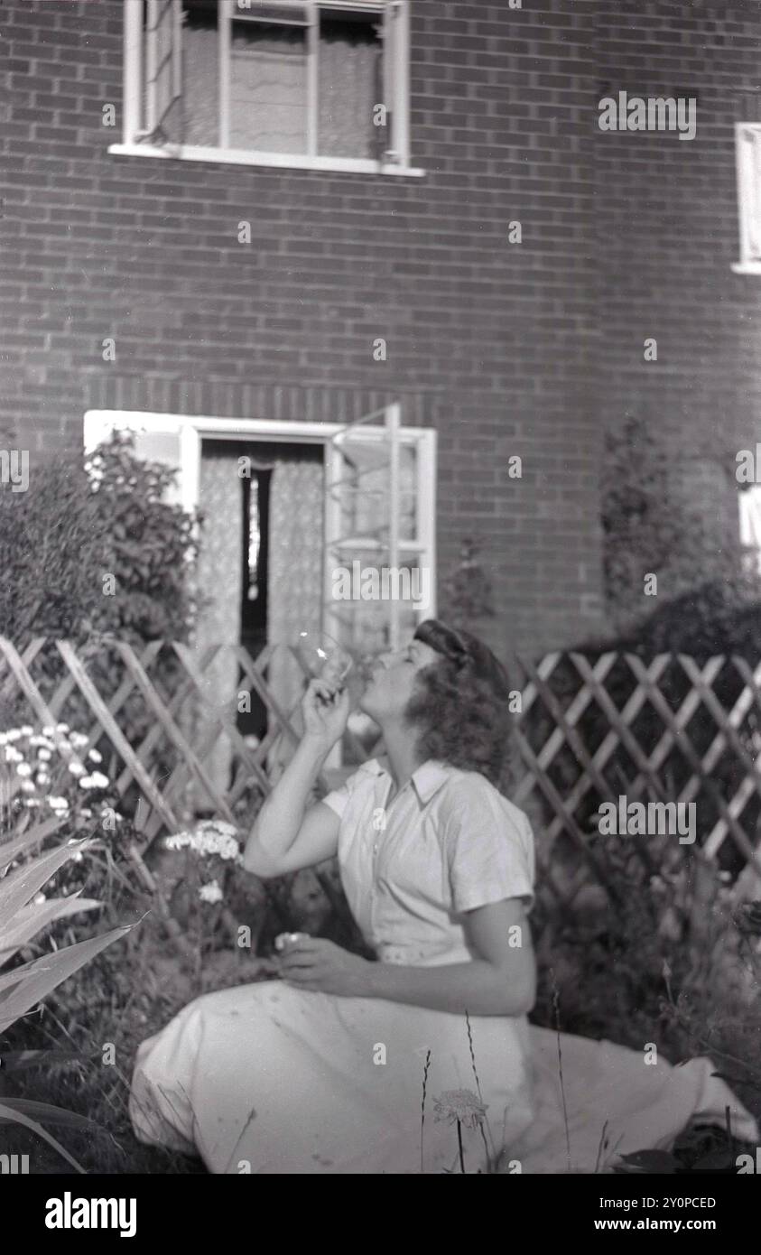 1960, storica, estiva e una giovane signora, seduta in un giardino sul retro di una casa di periferia, che soffia bolle attraverso un vetro, Inghilterra, Regno Unito. Foto Stock