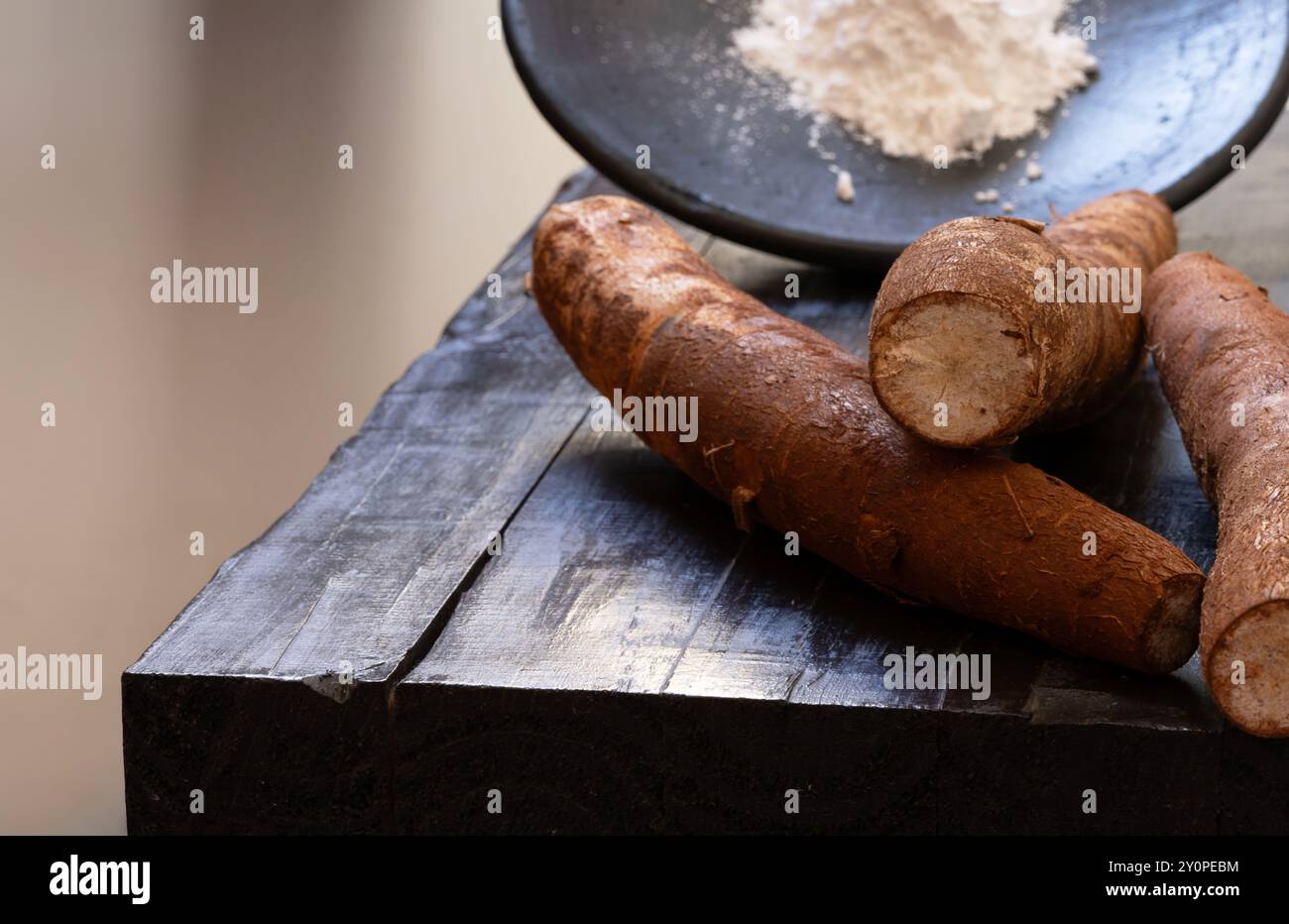 Tre radici di manioca con buccia sopra un tavolo di legno scuro con un piatto di farina di tapioca sullo sfondo fuori fuoco. Foto Stock