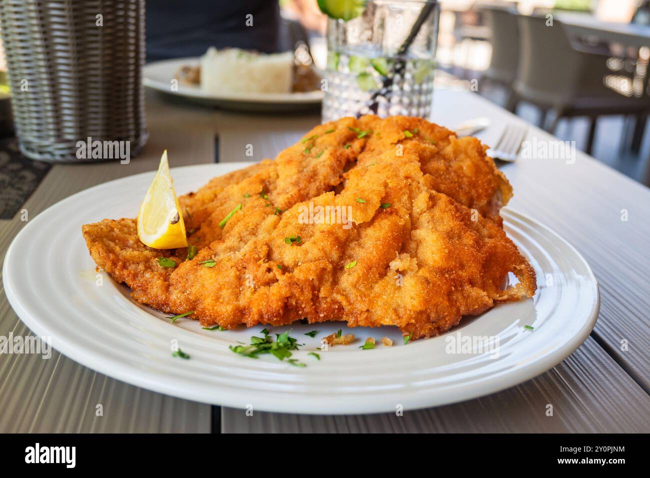 Schnitzel viennese su piatto bianco con bicchiere di bevanda, secondo piatto sul tavolo nel ristorante all'aperto. Foto Stock