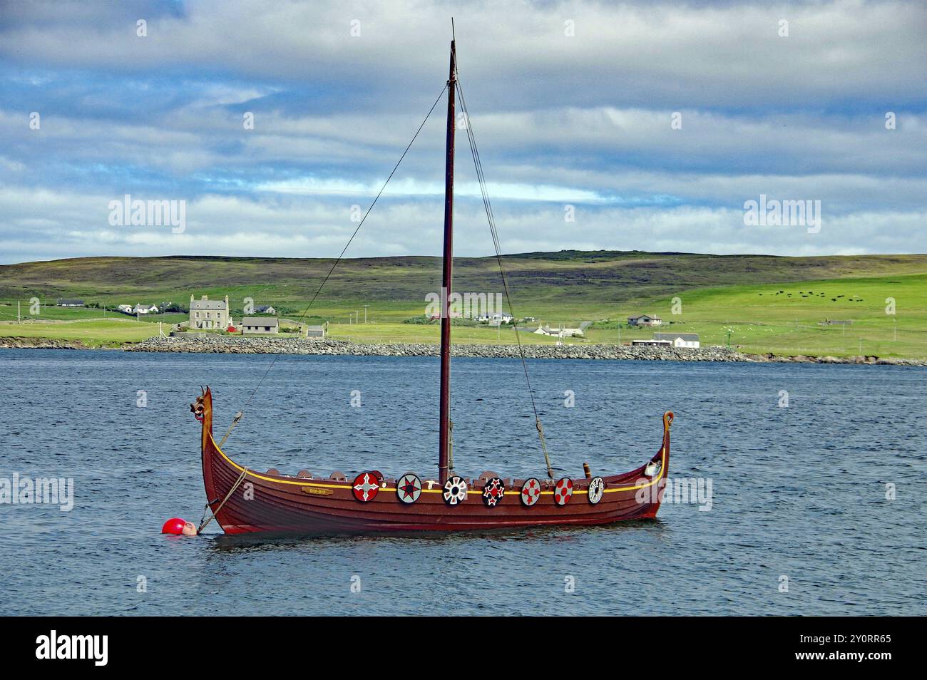 Una tradizionale barca vichinga con scudi sulle acque calme, che si affaccia su una costa verde e colline sullo sfondo, fino a Helly A, Lerwick, Shetland ISL Foto Stock