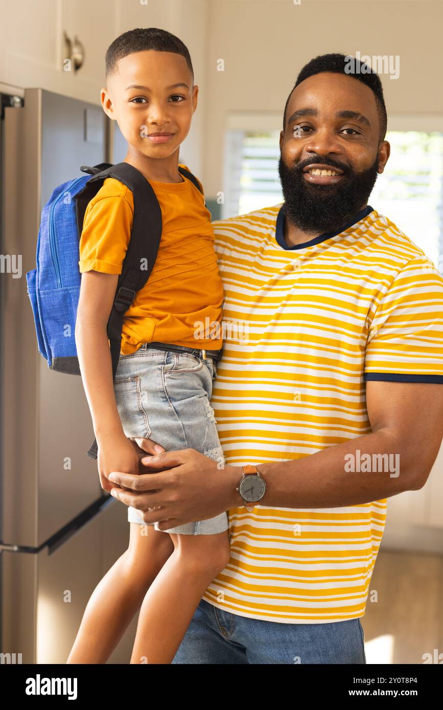 Padre che tiene il figlio con lo zaino, sorridente e pronto per la scuola in cucina Foto Stock
