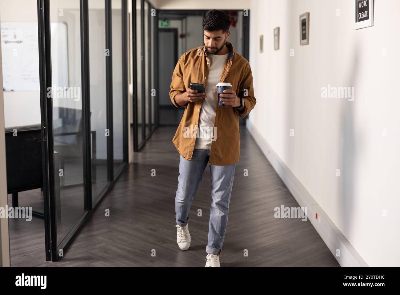 Cammina in ufficio, uomo indiano che usa lo smartphone e tiene una tazza di caffè Foto Stock