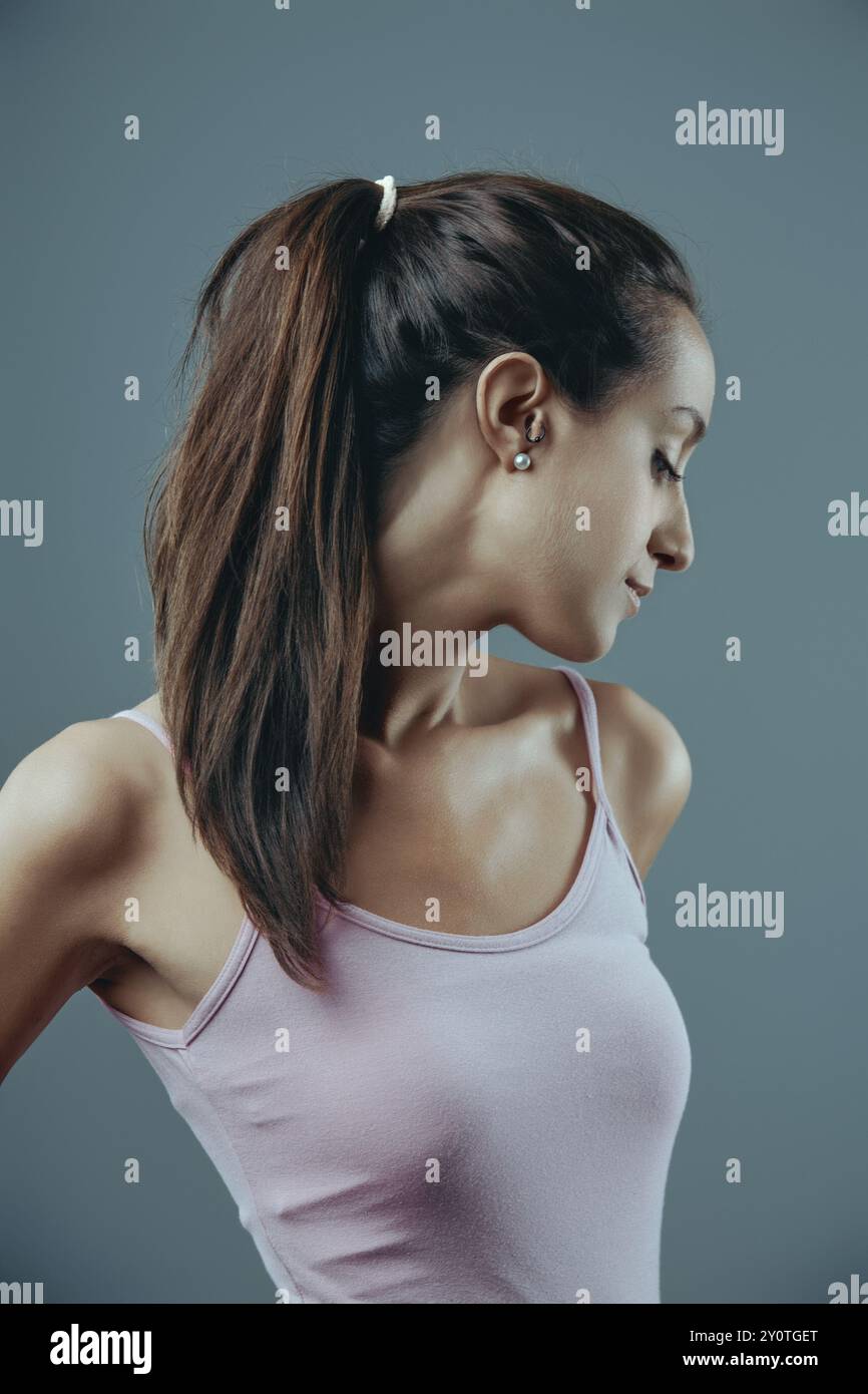La giovane donna sta posando con la testa inclinata verso il basso e un'espressione premurosa sul viso. Indossa una canotta rosa e i capelli mi vengono tirati indietro Foto Stock