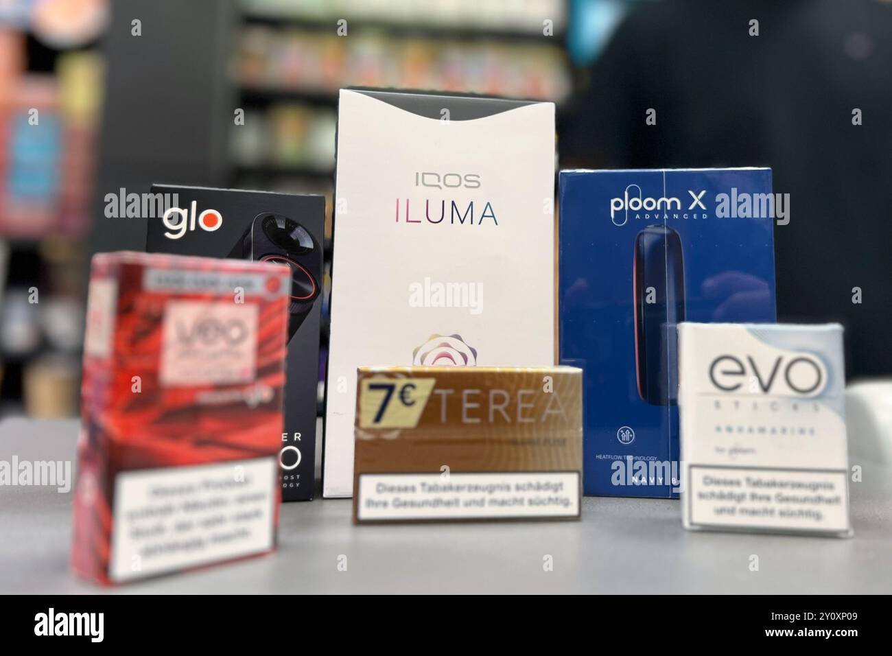 Duesseldorf, Germania. 4 settembre 2024. I riscaldatori elettrici per tabacco dei marchi Glo di British American Tobacco (BAT, sinistra), Iqos di Philip Morris (centro) e Ploom di Japan Tobacco International (JTI, destra) con i marchi corrispondenti di bastoncini per tabacco si trovano al banco di vendita di un negozio di tabacco, con un venditore alle spalle. Le grandi aziende del tabacco stanno investendo miliardi in riscaldatori per tabacco, in cui il tabacco non viene più bruciato ma semplicemente riscaldato. Essi posizionano i prodotti come un'alternativa meno dannosa alle sigarette tradizionali. Crediti: Wolf/dpa/Alamy Live News Foto Stock