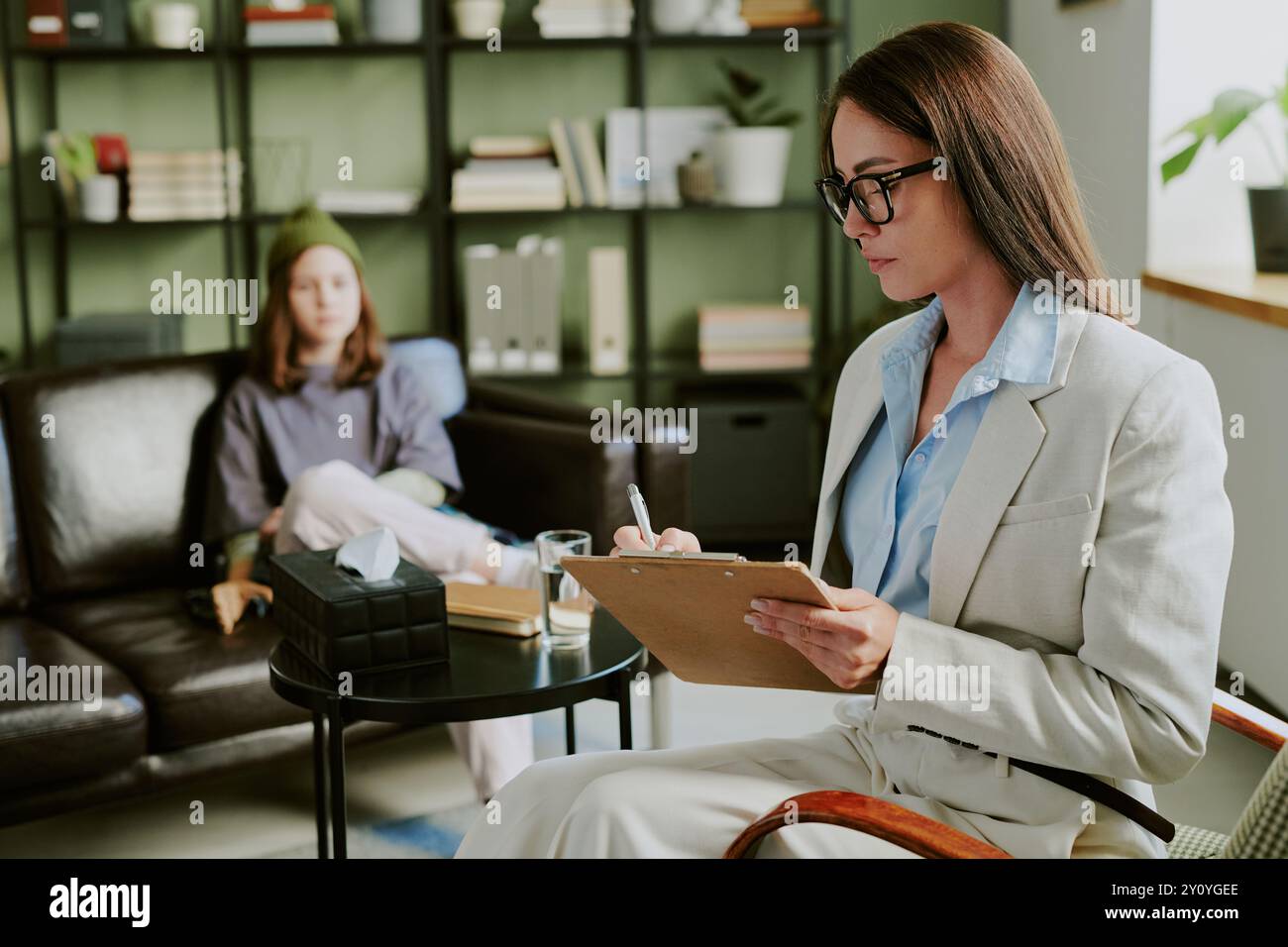 Consulente professionista che conduce una sessione tenendo gli appunti, cliente seduto sul divano con un atteggiamento premuroso. Interno con libreria organizzata A. Foto Stock