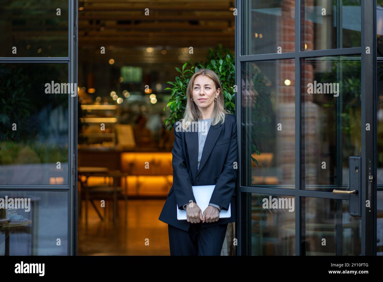 Donna premurosa e sicura di sé con un portatile in mano all'ingresso del caffè durante la mattinata a riflettere. Foto Stock