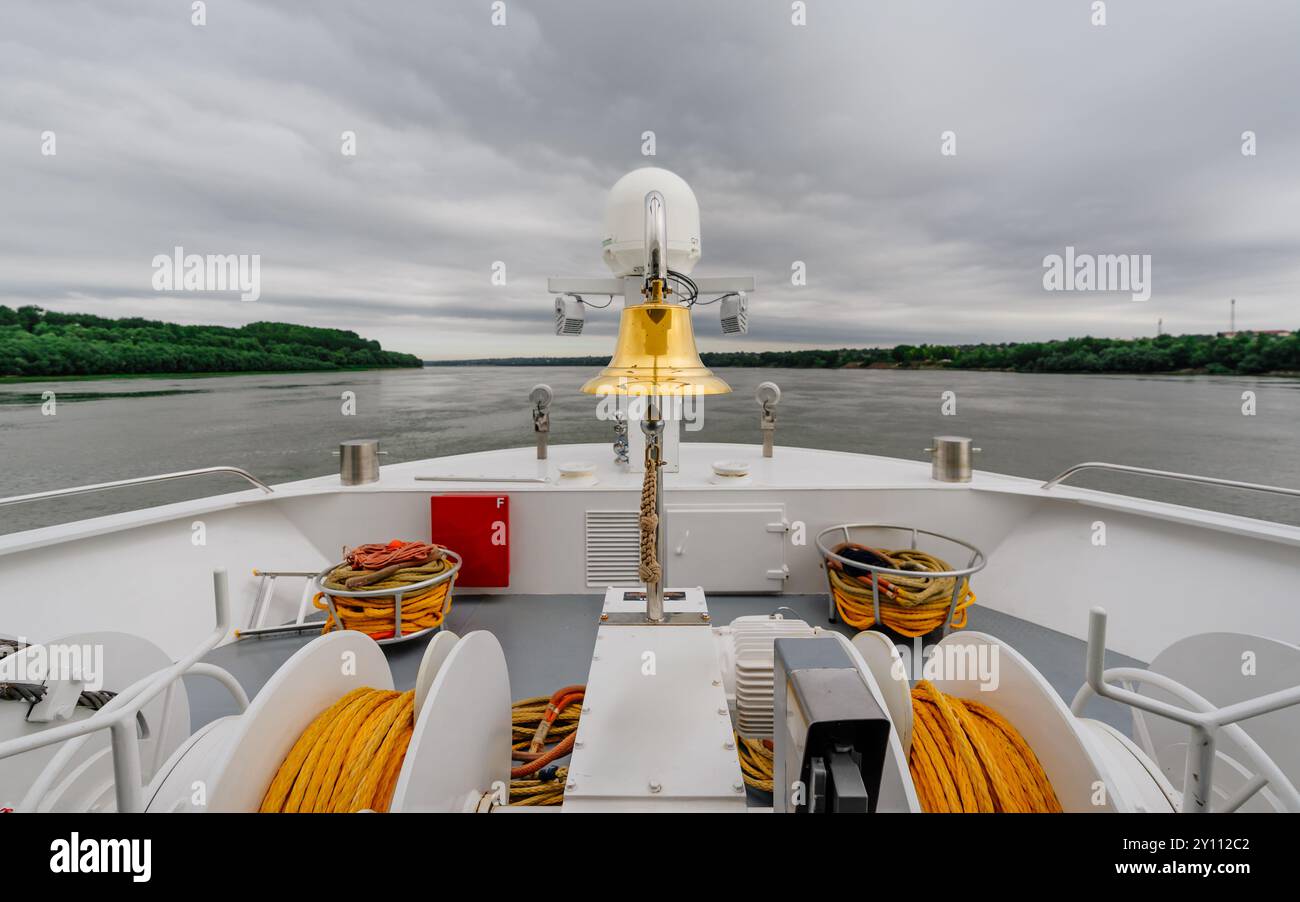 Prua della nave con equipaggiamento di navigazione, corde e campana che si affaccia sul fiume e sulla costa boscosa Foto Stock