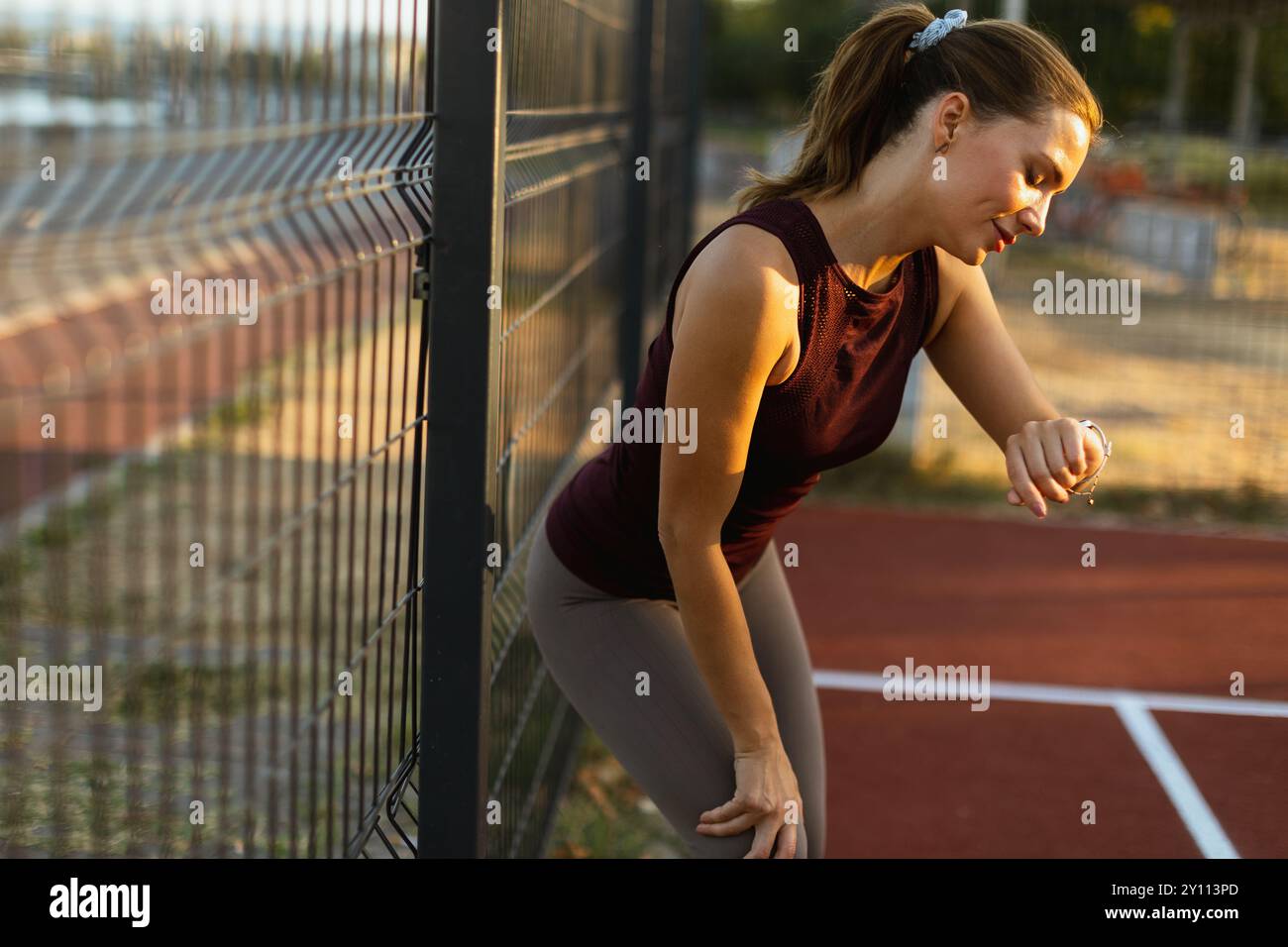 Esausta ma determinata, una donna monitora il suo tempo mentre si appoggia contro una recinzione dopo un'intensa sessione di corsa su una pista vivace durante il Golden Foto Stock
