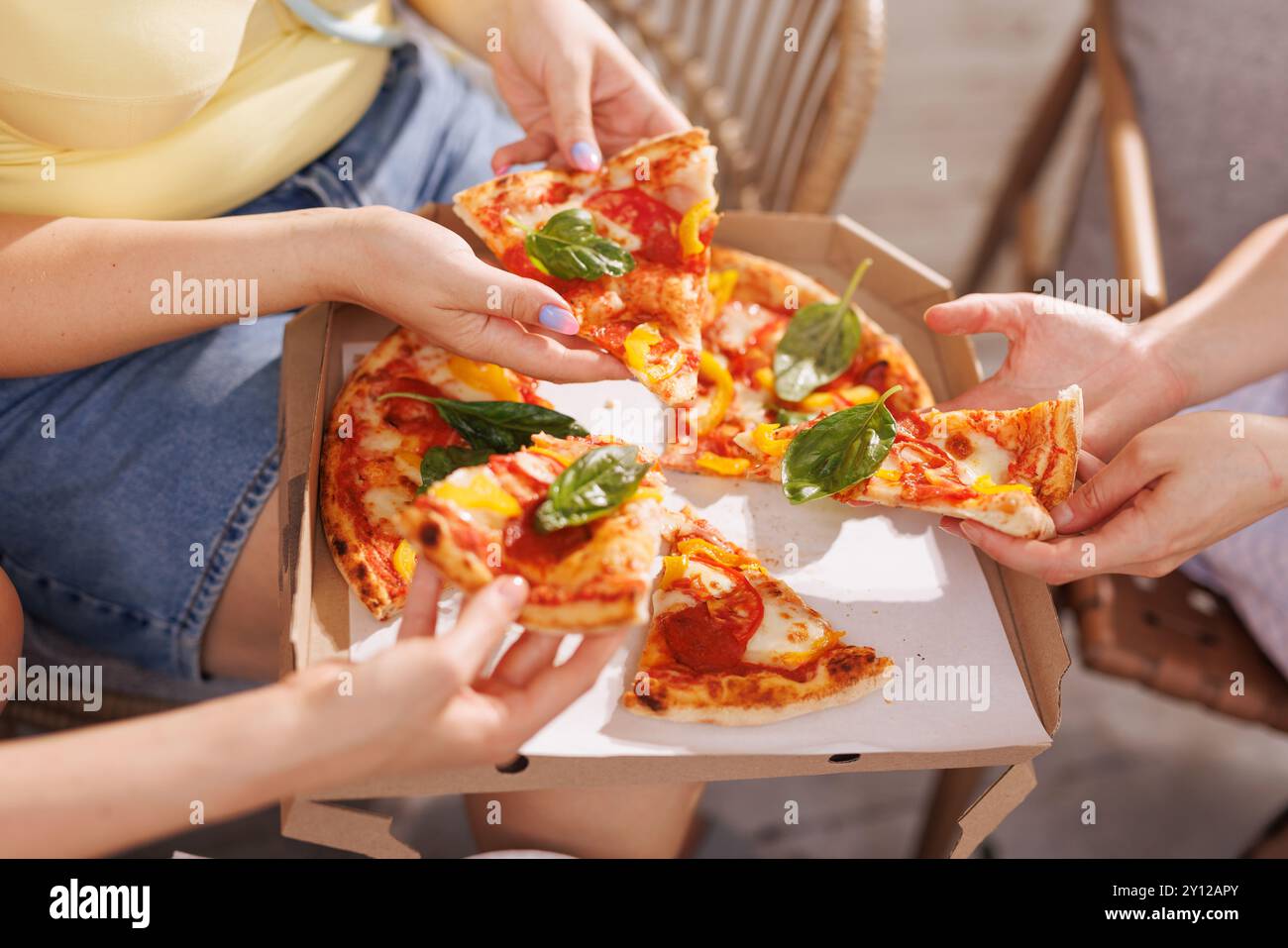 Un gruppo di amici condivide la pizza all'aperto, le mani si avvicinano per le fette guarnite con basilico fresco, mentre una scatola per pizza e piatti di antipasti si siedono sulla schiena Foto Stock