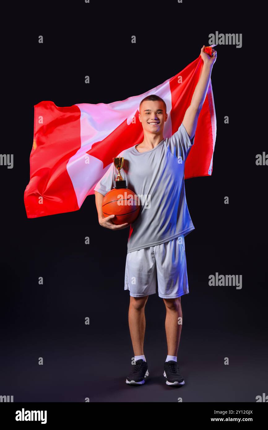 Giovane giocatore di basket maschile con bandiera canadese, coppa d'oro e palla su sfondo nero Foto Stock