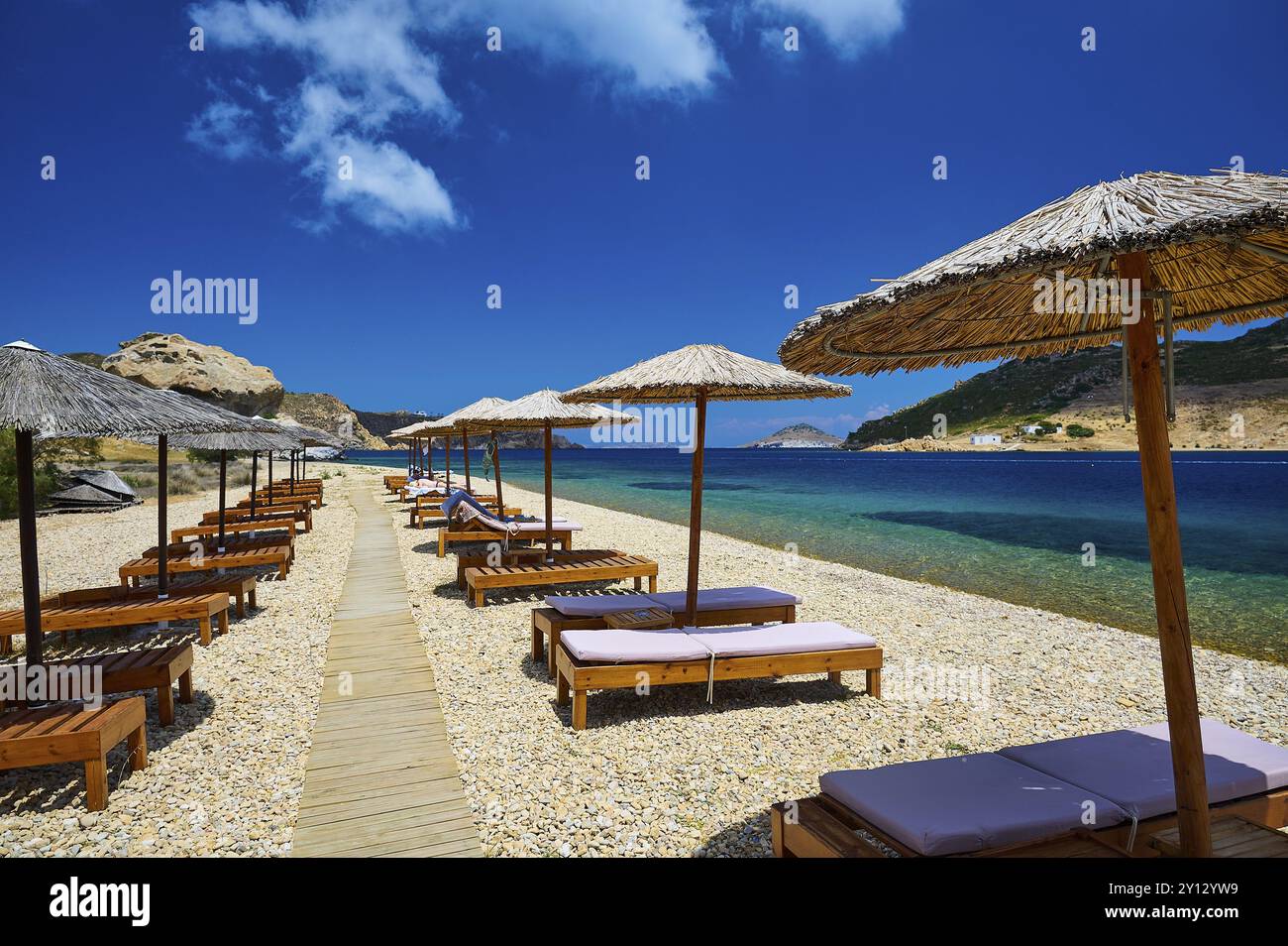 Sdraio e ombrelloni sulla spiaggia con passerella in legno, vista libera del mare calmo e del cielo blu, Petras Beach, Kalikatsou Rock, Patmos, Dodecaneso, Foto Stock