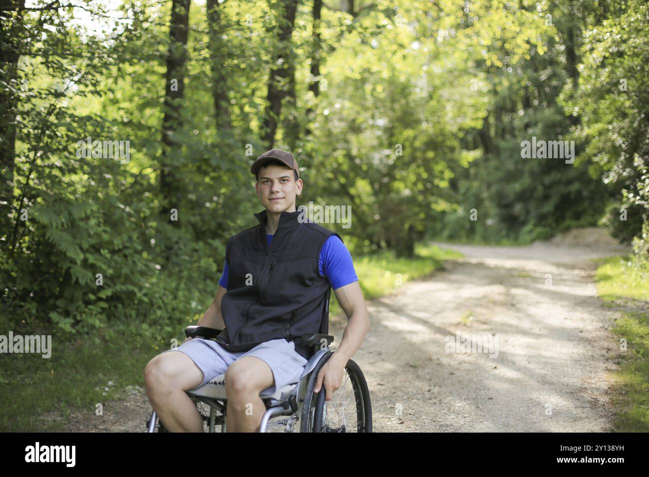 Felice e giovani disabili o handicappati uomo seduto su una sedia a rotelle in natura girando le ruote su una strada a piedi in una bella giornata di sole Foto Stock