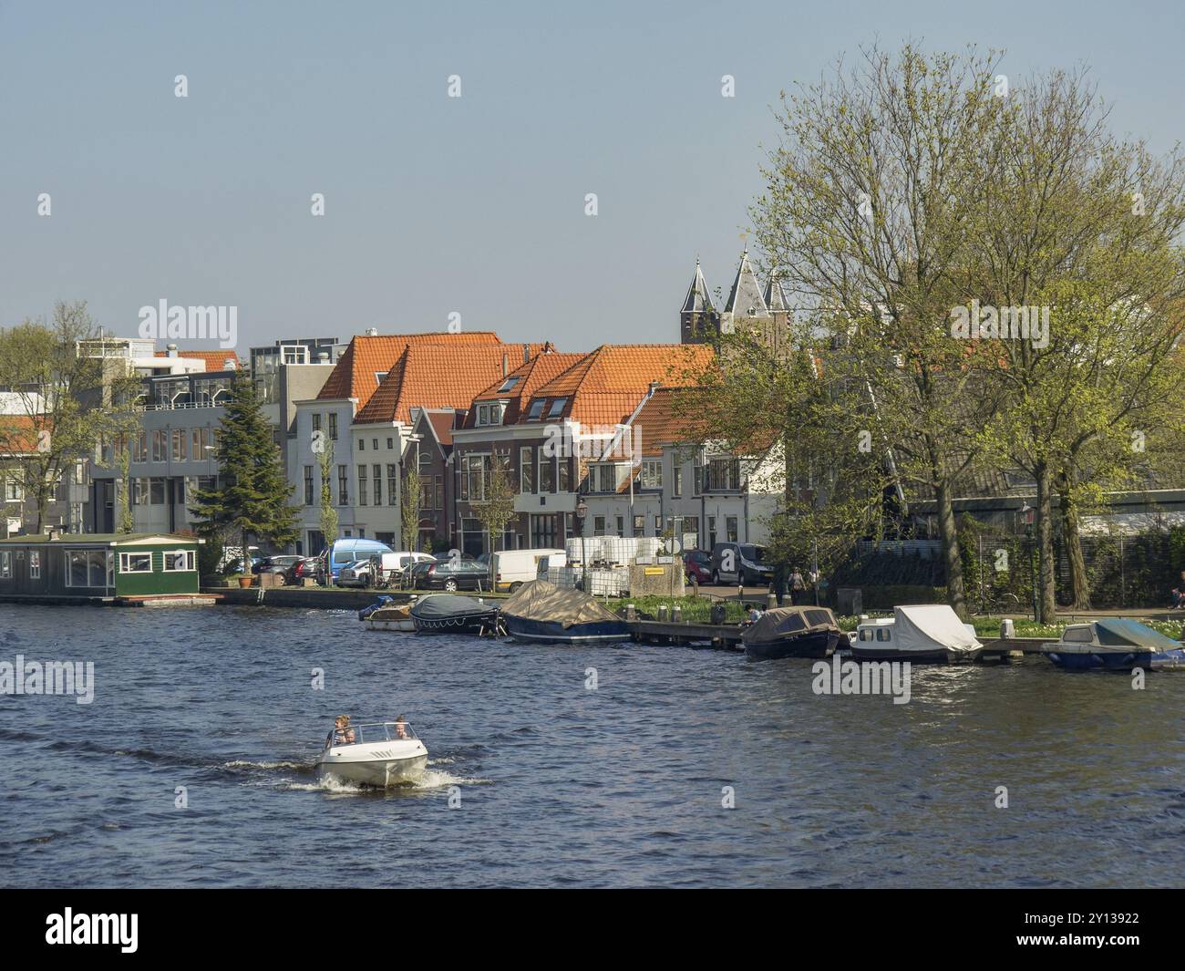 Naviga in barca su un fiume lungo una fila di case con tetti rossi, circondate da alberi verdi, sotto un cielo azzurro e limpido, Haarlem, Paesi Bassi Foto Stock