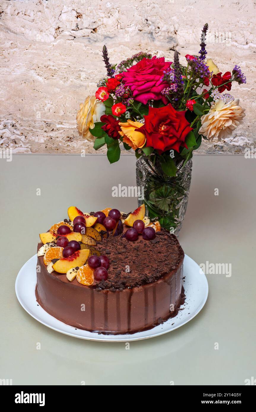 Una torta al cioccolato decorata con frutta, accanto a una vibrante composizione floreale in un vaso. Foto Stock