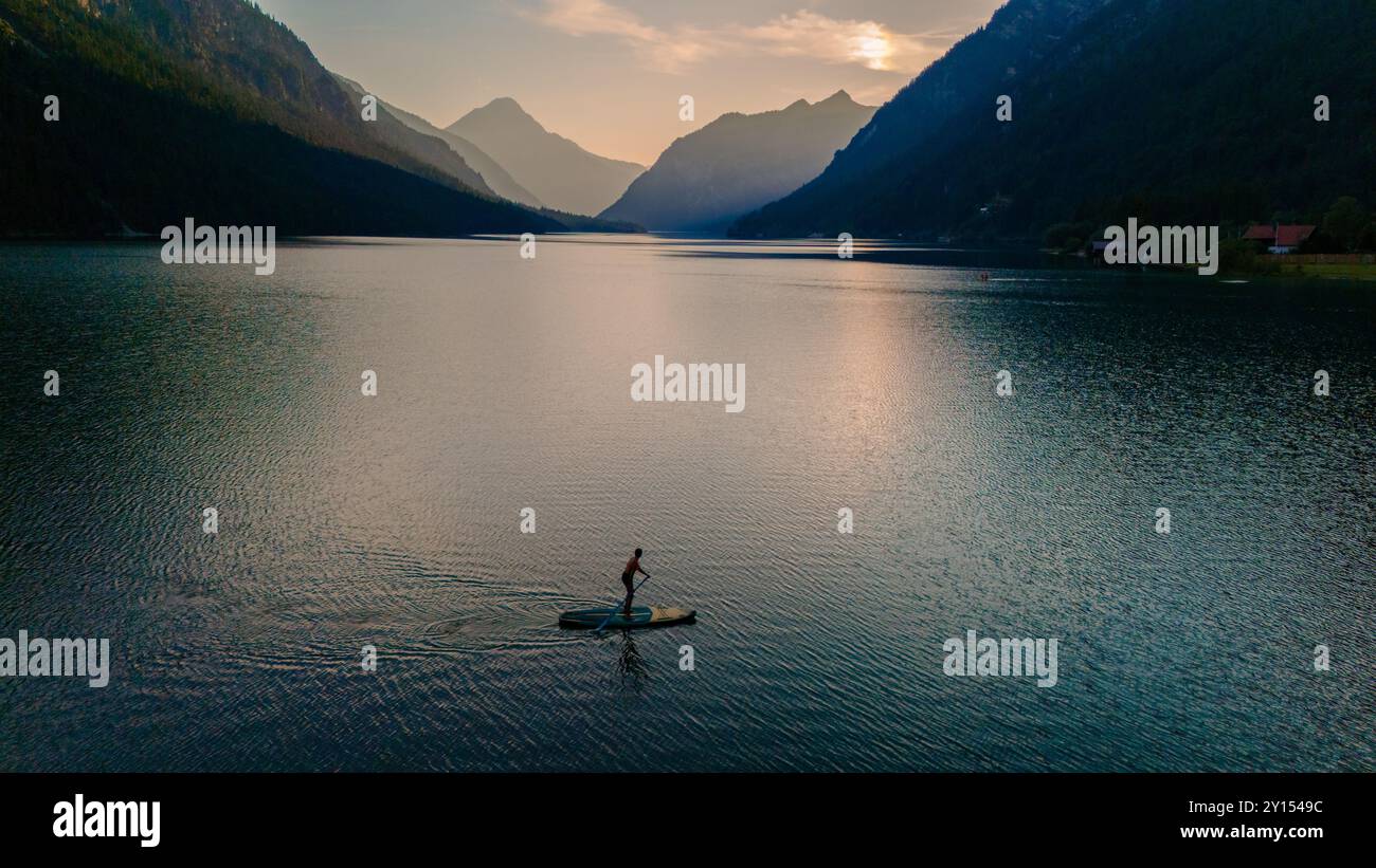 Mentre il sole scende sotto le montagne, un avventuriero solitario scivola attraverso le tranquille acque di Plansee, circondato dallo splendido paesaggio alpino e dal soffice bagliore del crepuscolo. Foto Stock