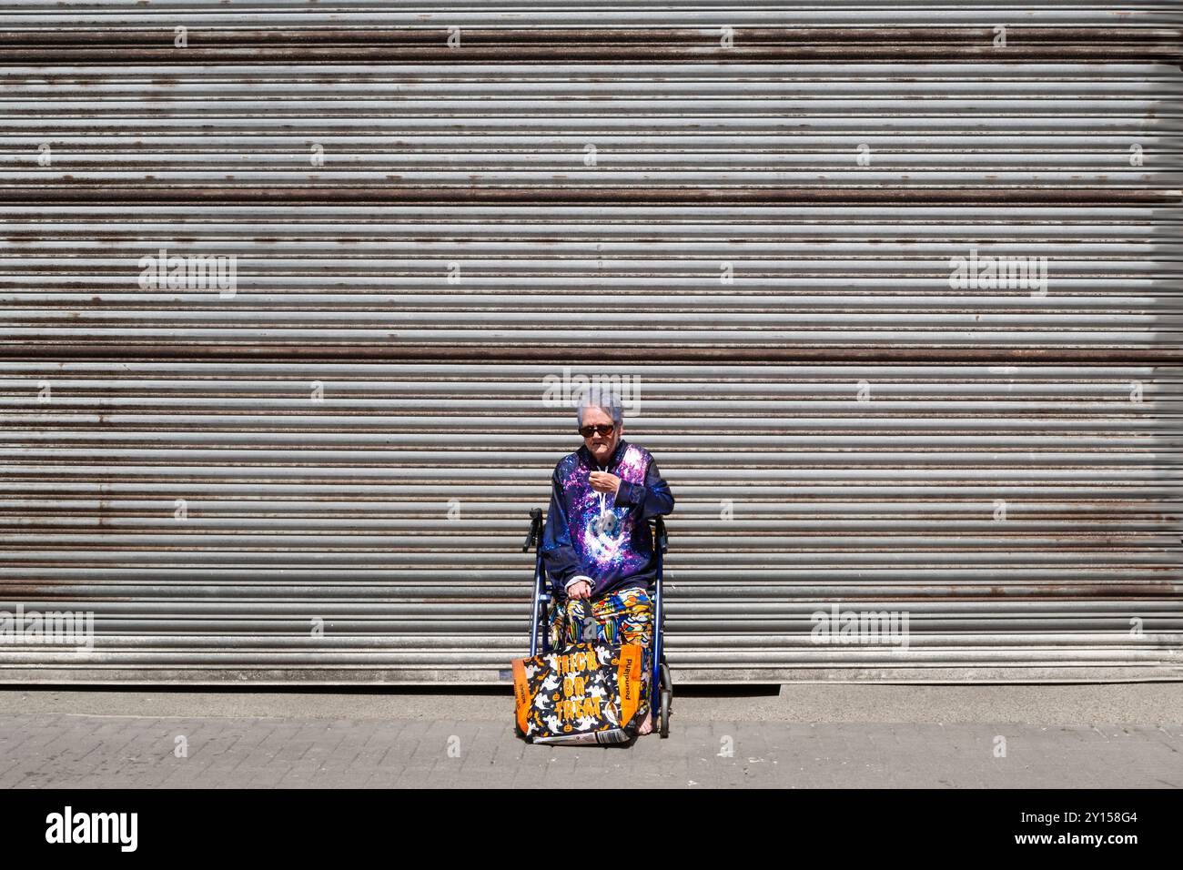 Una donna che indossa abiti dai colori vivaci e sgargianti seduti da sola su un telaio per camminare con sedia a rotelle, fuori dalle persiane di un negozio chiuso a Newquay T. Foto Stock