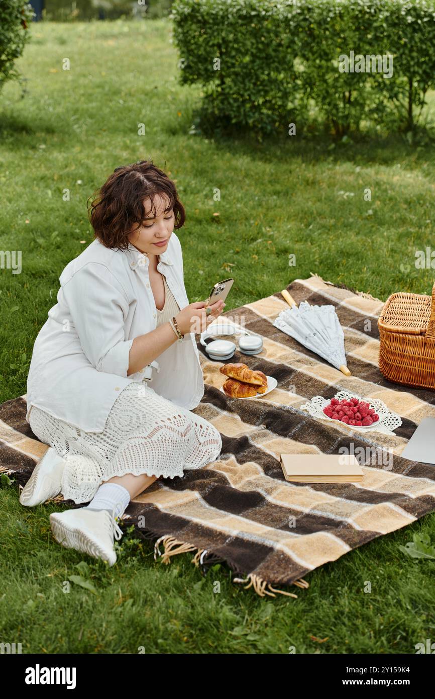 Una donna con una camicia bianca riposa su una coperta, assaporando un picnic estivo in un lussureggiante parco verde. Foto Stock