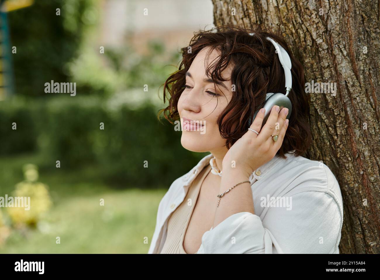 Una giovane donna con una camicia bianca si rilassa sotto un albero, immergendosi nel calore estivo mentre ascolta musica. Foto Stock