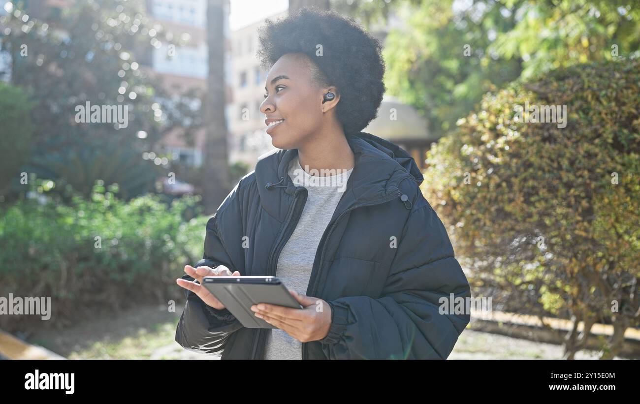 La donna afroamericana con i capelli ricci ama la musica tramite auricolari wireless mentre tiene in mano un tablet all'aperto. Foto Stock