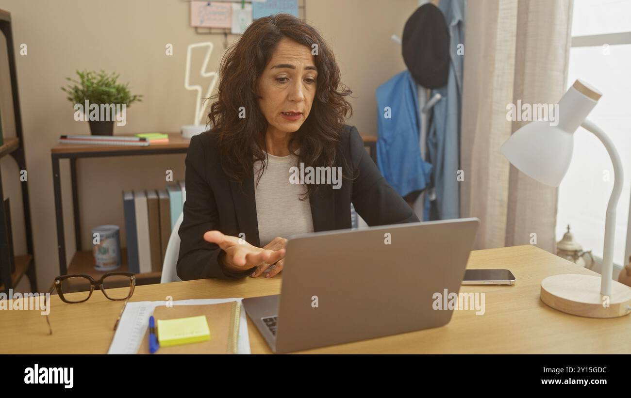 Una donna di mezza età partecipa a una videochiamata da un ufficio domestico interno, mostrando un comportamento casual ma professionale. Foto Stock