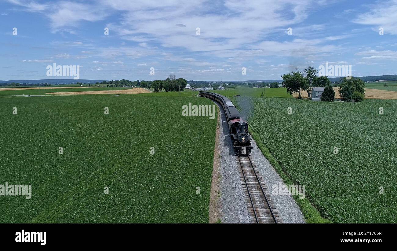 Una locomotiva a vapore d'epoca con vista aerea si muove lungo sentieri panoramici, circondati da ampi campi verdi e alberi torreggianti, mostrando il fascino dei paesaggi rurali in una giornata limpida. Foto Stock