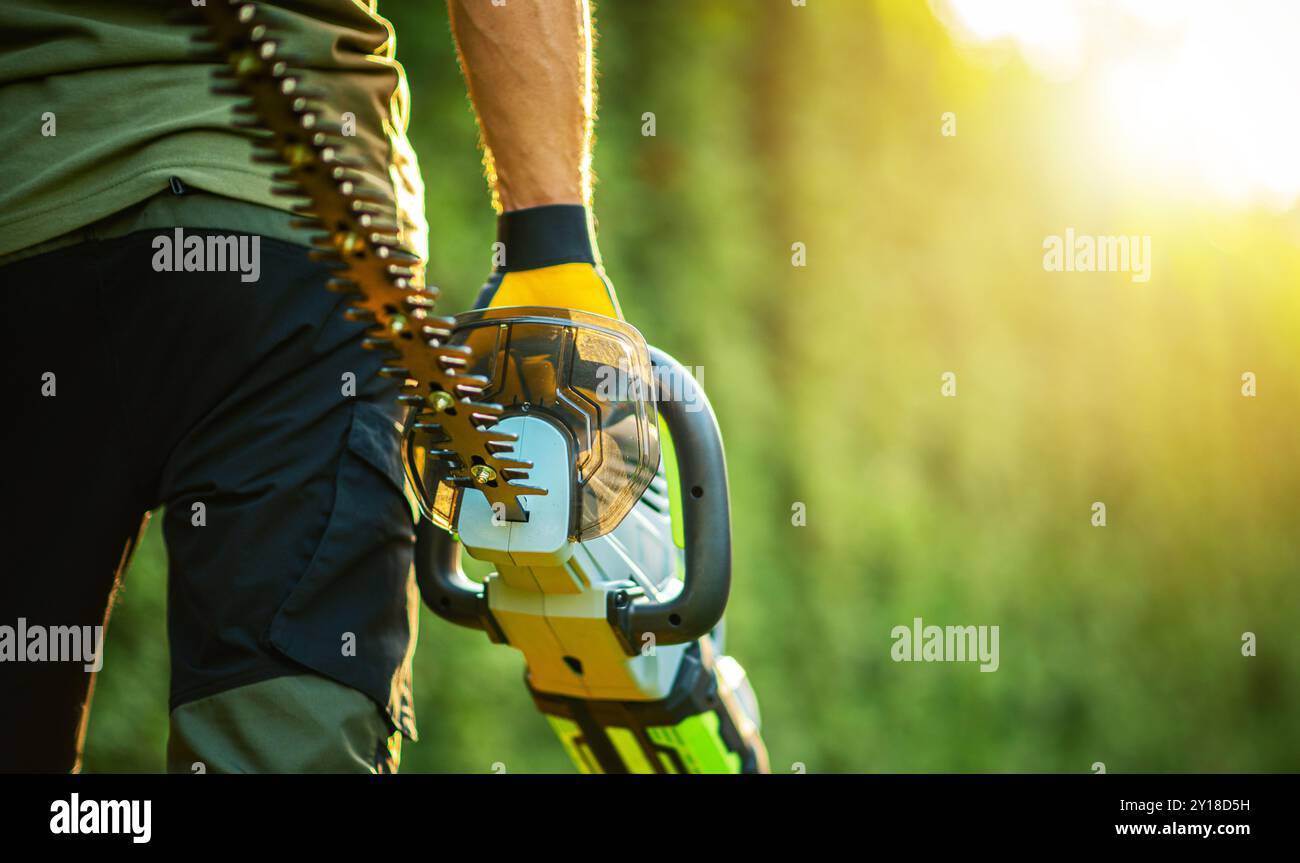 Un giardiniere si trova in un lussureggiante giardino verde con un tagliasiepi elettrico, pronto per rifinire gli arbusti durante il sole del pomeriggio estivo. Foto Stock