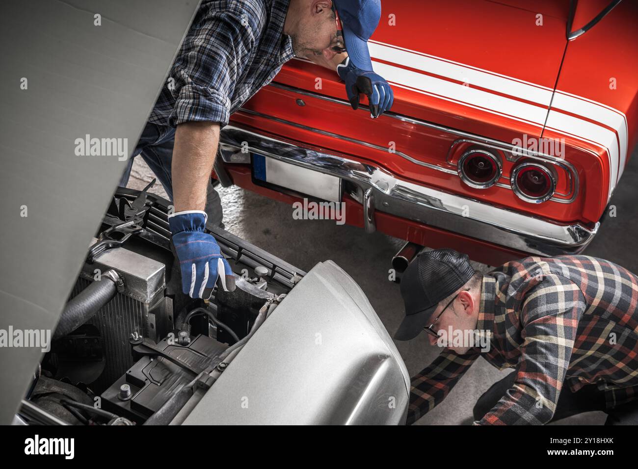 Due meccanici si concentrano sulla riparazione di un'auto d'epoca in un garage, mentre un'altra auto d'epoca è parcheggiata nelle vicinanze, mostrando il loro lavoro di squadra e la loro abilità. Foto Stock