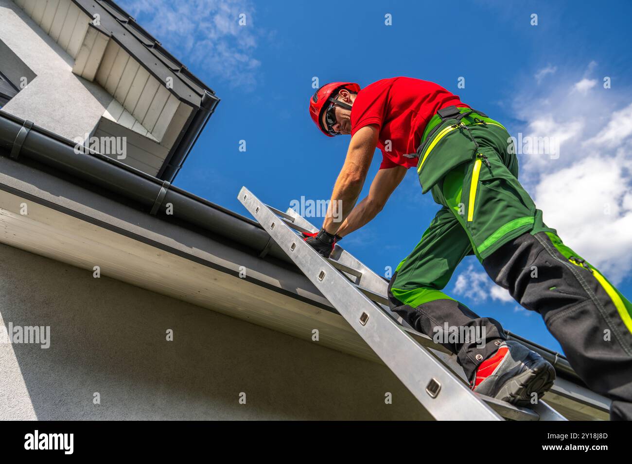 Un addetto alla manutenzione con l'attrezzatura di sicurezza sale su una scala per ispezionare e riparare una grondaia del tetto mentre il sole splende in alto. Foto Stock