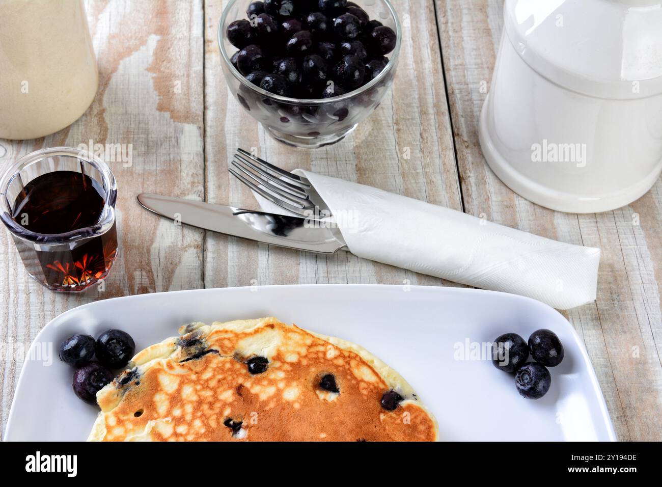 Primo piano di una pila di pancake di mirtilli freschi fatti in casa su un piatto bianco. Utensili e stoviglie per sciroppo d'acero e un tavolo in legno rustico. Foto Stock