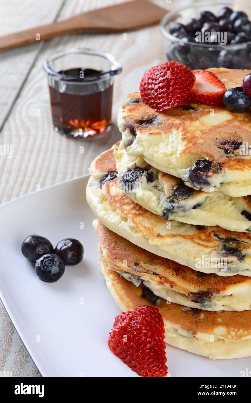 Primo piano di una colazione fatta in casa con pancake ai mirtilli con caraffa di sciroppo, spatola di legno, ciotola di mirtilli e fragole. Formato verticale con de basso Foto Stock