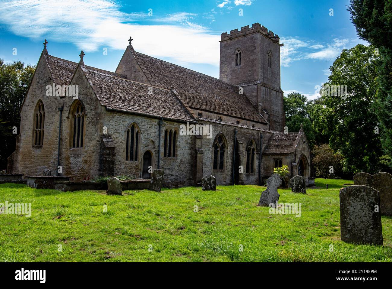 Storica chiesa in pietra della Santa Croce Ashton Keynes con un'alta torre, circondata da un lussureggiante cimitero verde, sotto un cielo blu parzialmente nuvoloso. Foto Stock