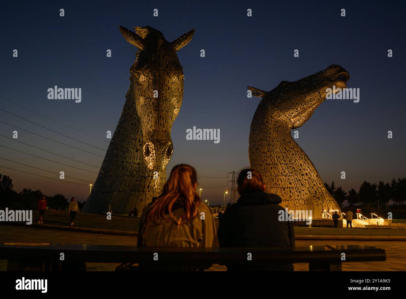 Due ragazze adorano la tranquillità al tramonto della più grande scultura equina del mondo, le iconiche Kelpies, all'Helix Park, Falkirk, Scozia, REGNO UNITO Foto Stock