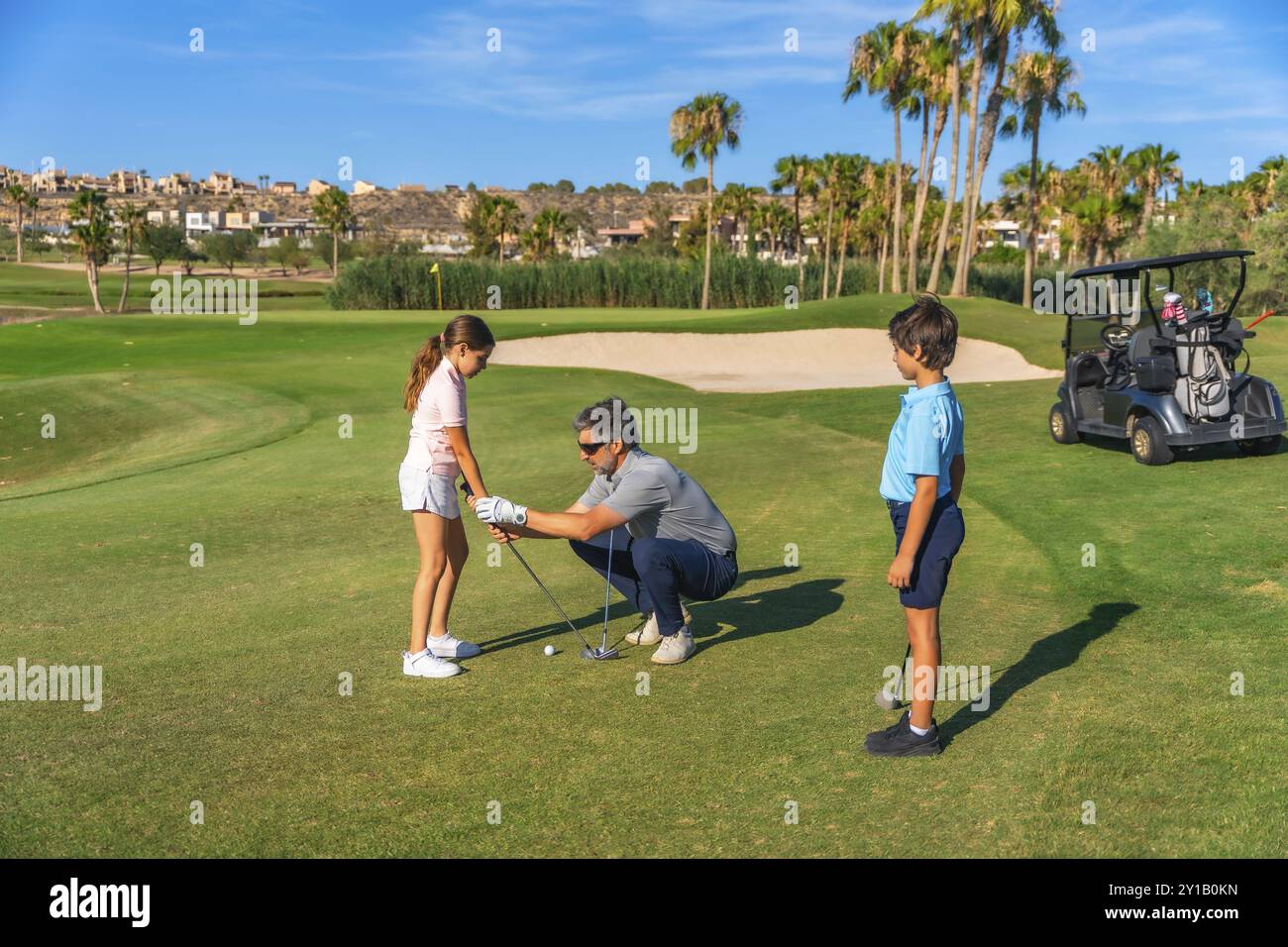 Istruttore maturo maschio che insegna a una ragazza come giocare a golf accanto a un ragazzo in un lussuoso campo verde oro Foto Stock