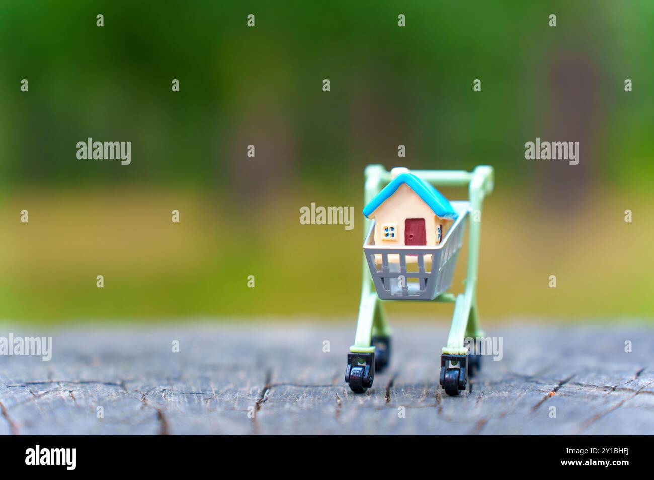 Carrello della spesa in miniatura che porta un modello di piccola casa, posizionato su una superficie rustica in legno, simboleggia gli immobili, l'acquisto di case e una divertente truffa finanziaria Foto Stock