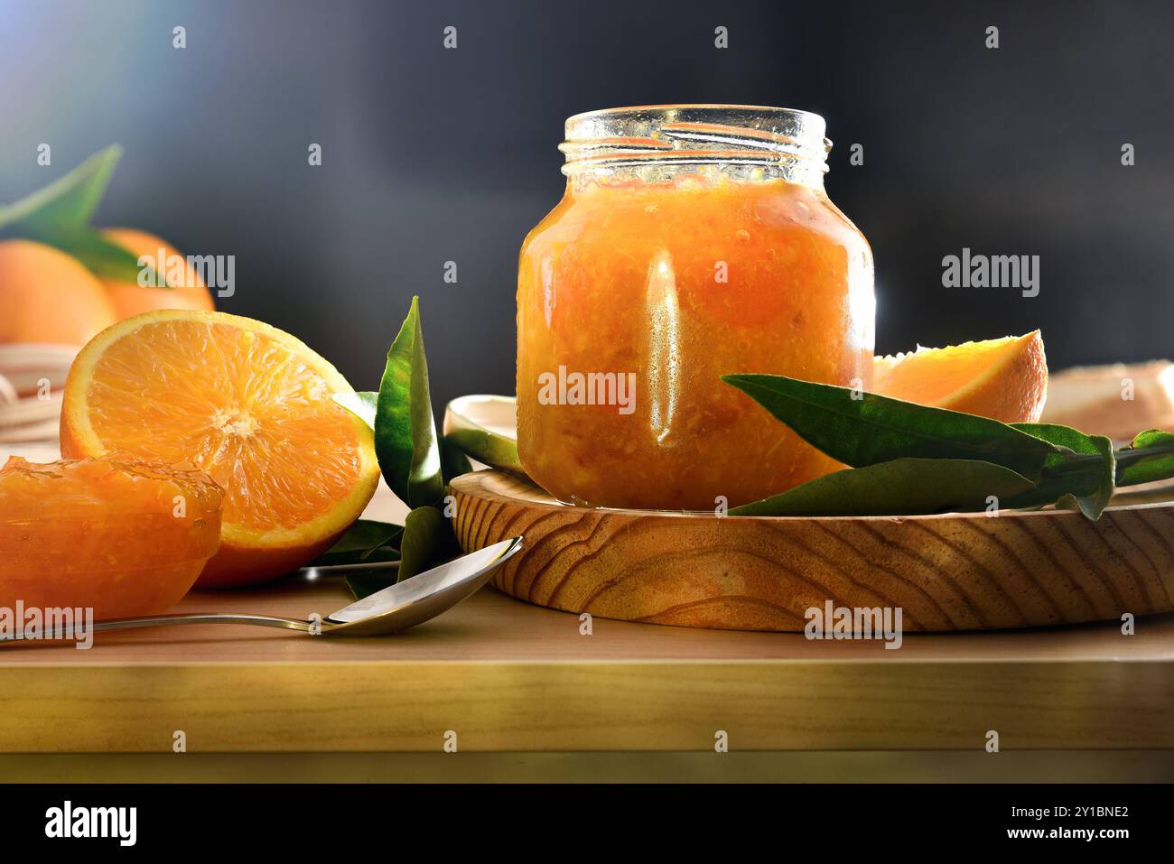 Dettaglio del vaso con marmellata d'arancia e tagliare la frutta intorno ad esso su banco da cucina in legno e sfondo scuro. Vista frontale. Foto Stock