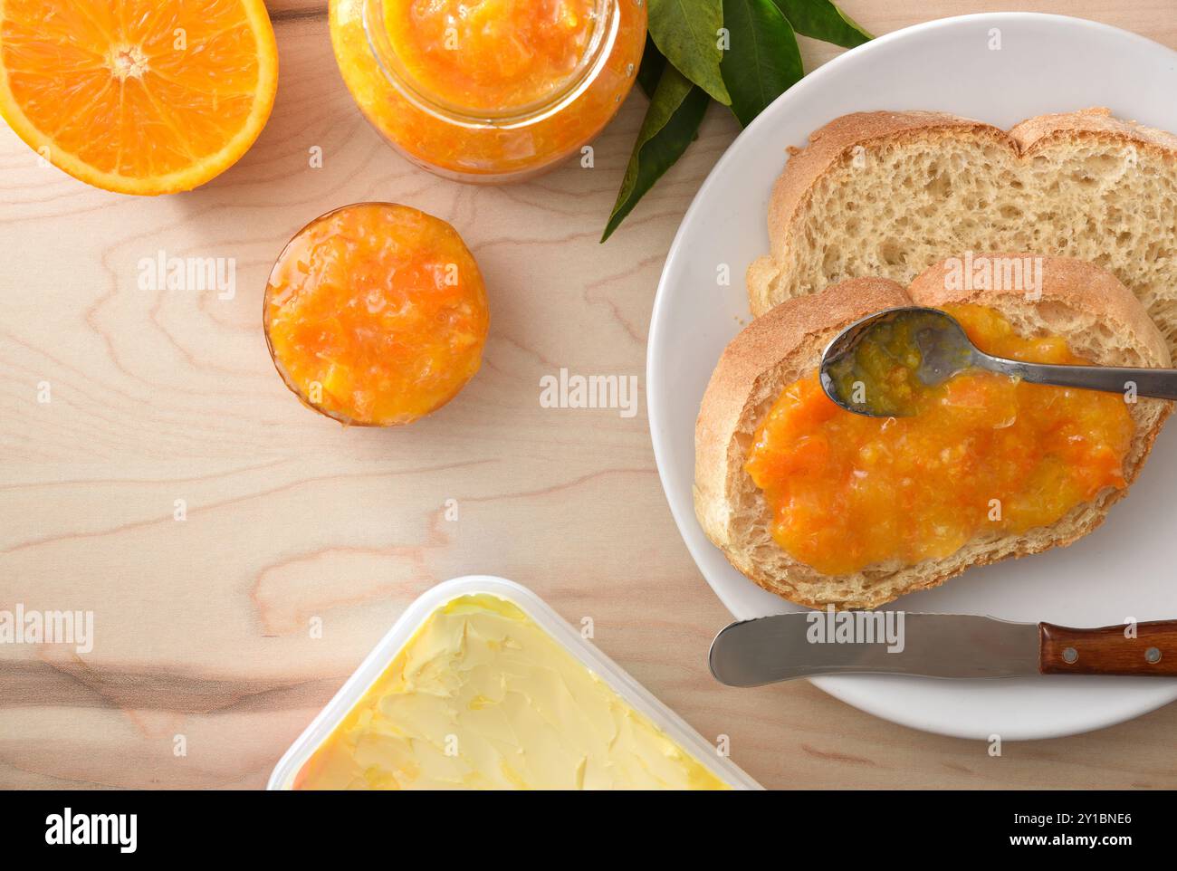 Dettaglio della marmellata d'arancia spalmata su una fetta di pane e in contenitori di vetro su una tavola di legno con burro e posate. Vista dall'alto. Foto Stock