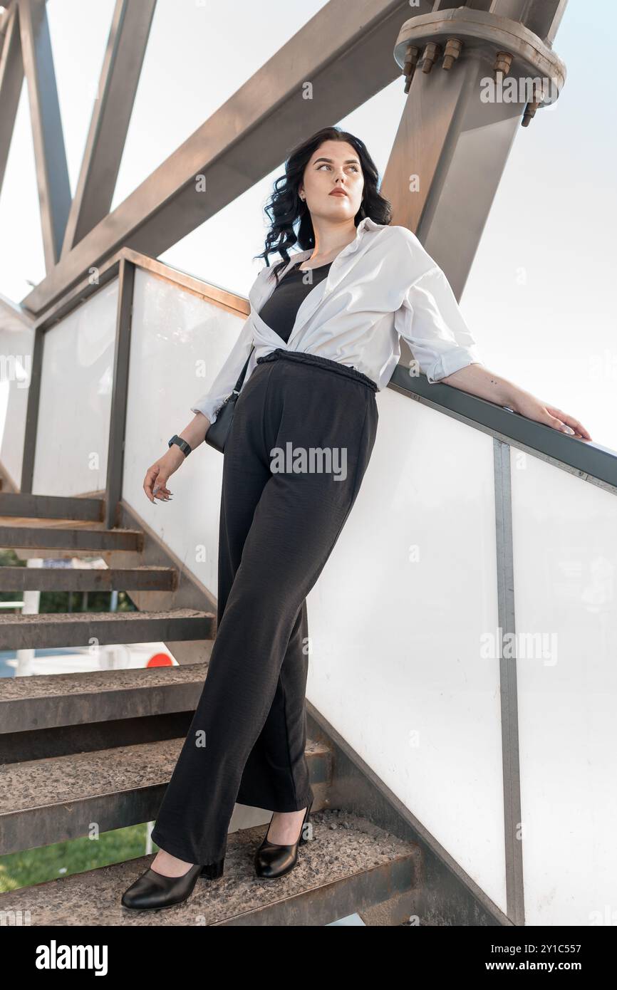Donna sicura di sé in un abbigliamento professionale che posa sulla scala industriale sotto l'architettura moderna. Foto Stock