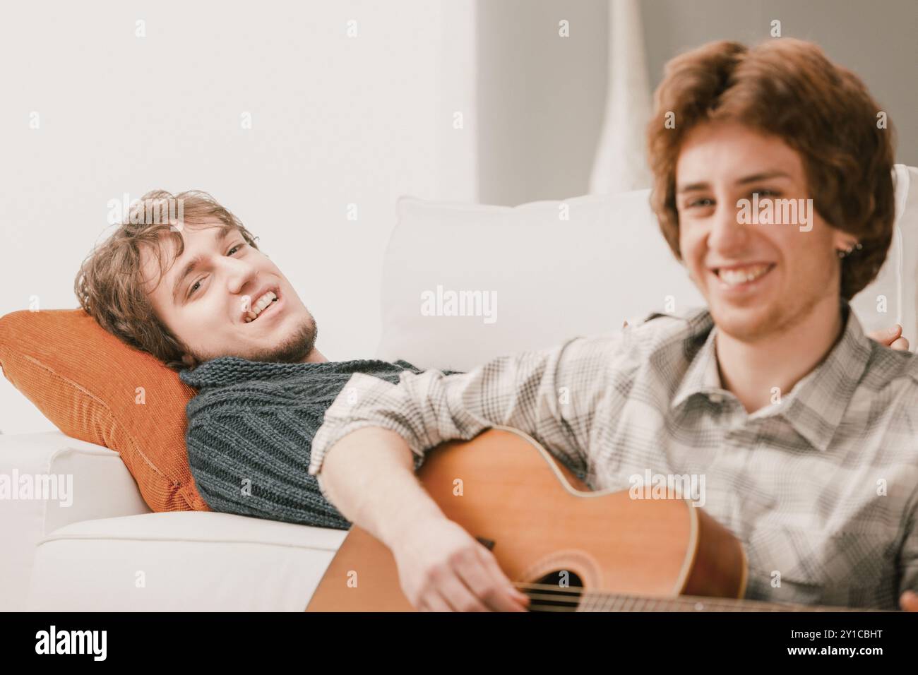 Due amici che si rilassano su un divano, suonano la chitarra, ridono e si godono il comfort di casa. Condivisione di musica e gioia in un soggiorno moderno. Tempo di qualità con Foto Stock