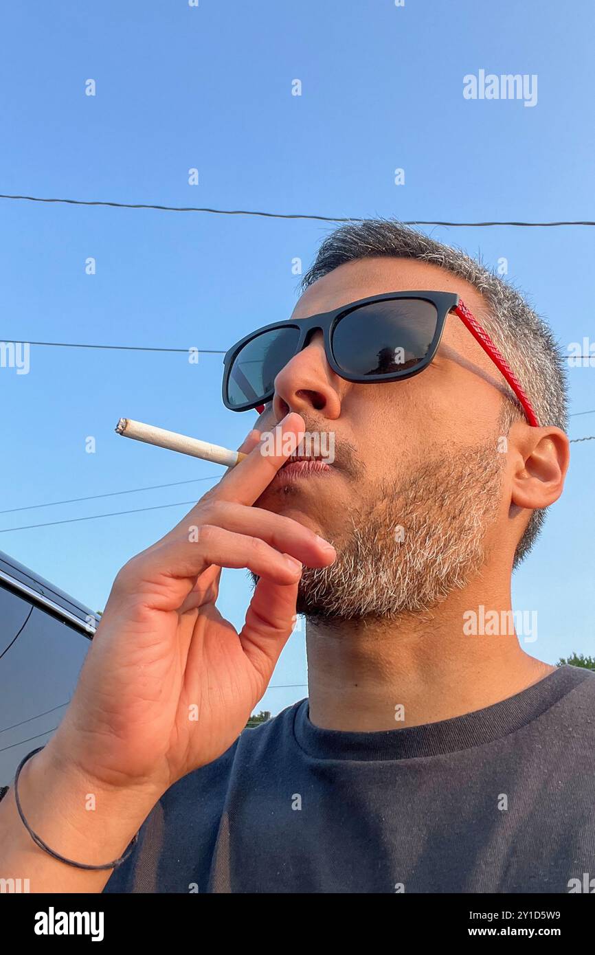 Un uomo sta fumando una sigaretta mentre indossa gli occhiali da sole. Concetto di relax e svago, mentre l'uomo si prende una pausa dalla sua giornata per gustare un sigaretto Foto Stock Un uomo sta fumando una sigaretta mentre indossa gli occhiali da sole. Concetto di relax e svago, mentre l'uomo si prende una pausa dalla sua giornata per gustare un sigaretto Foto Stock