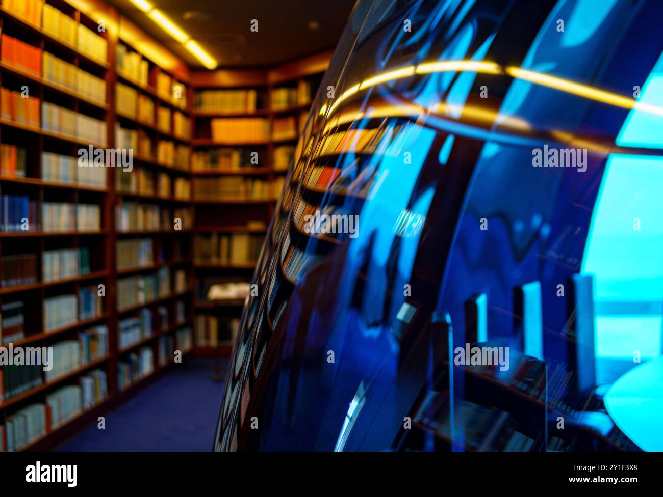 Una cattura artistica di una moderna biblioteca con scaffali pieni di libri, riflessa in una superficie curva in vetro di colore blu Foto Stock