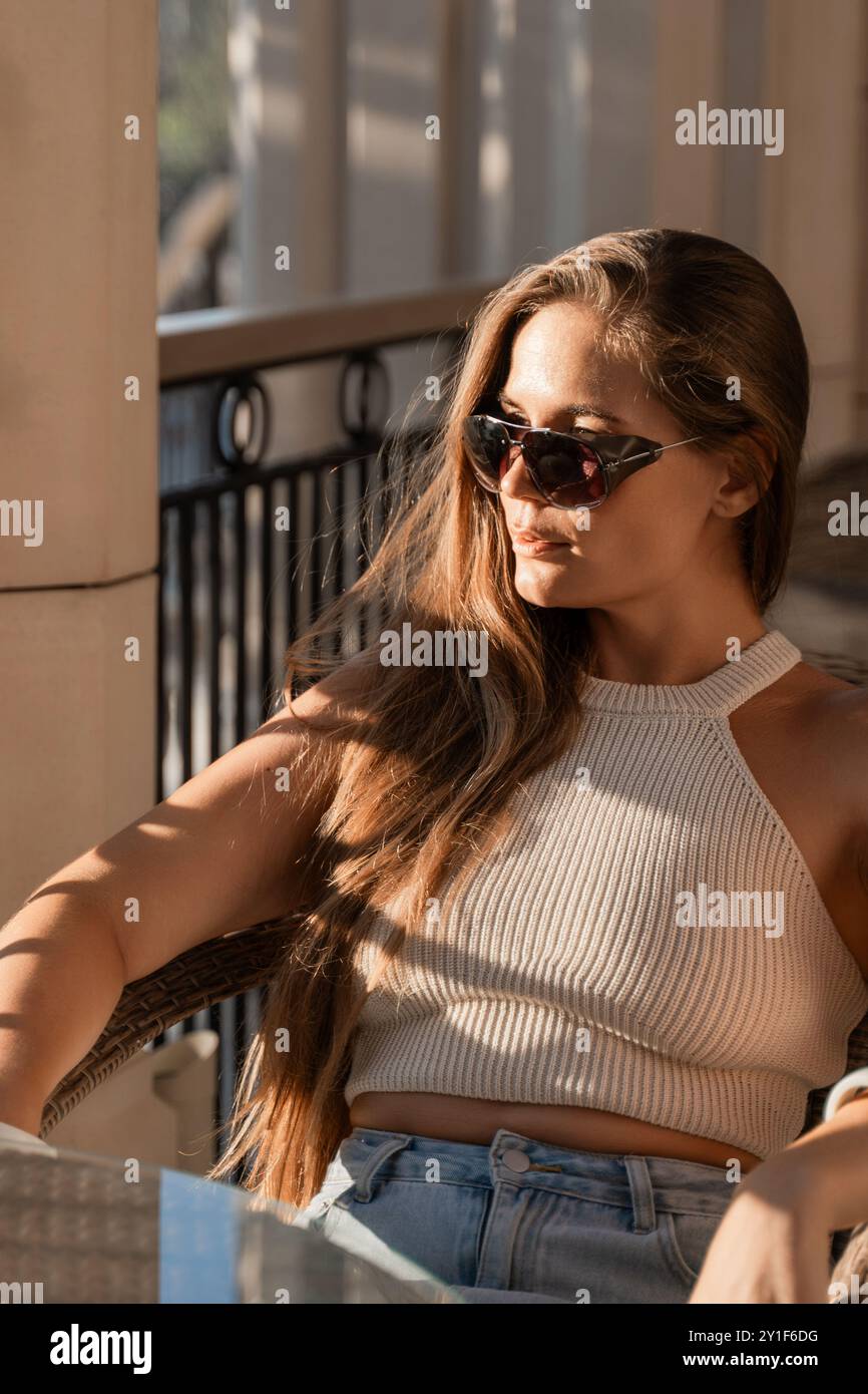 Una donna con lunghi capelli marroni, che indossa occhiali da sole e una canotta bianca, siede su un patio e guarda lateralmente. Foto Stock