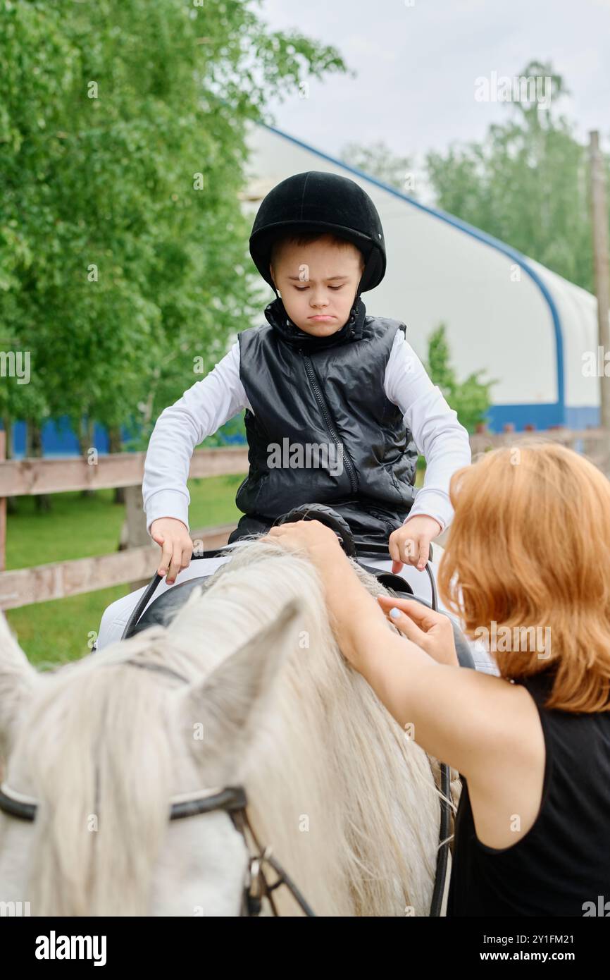 Bambino seduto su un cavallo bianco mentre viene assistito da una donna adulta in un ambiente all'aperto in una giornata nuvolosa circondato da alberi e strutture Foto Stock