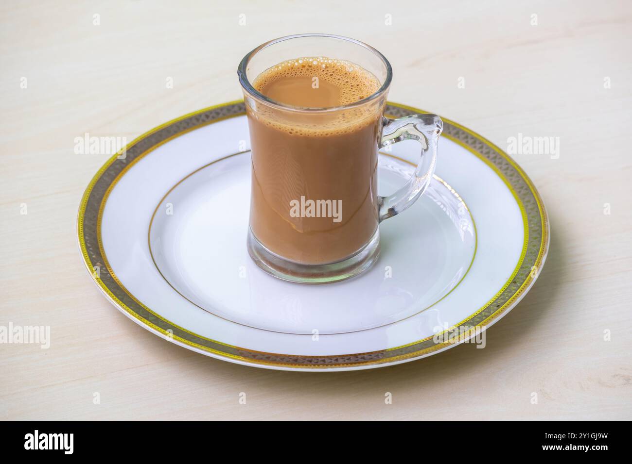 Una tazza di tè al latte caldo o di dudh cha in una tazza di bicchiere, servita in un piatto bianco su una superficie di legno chiaro. Foto Stock