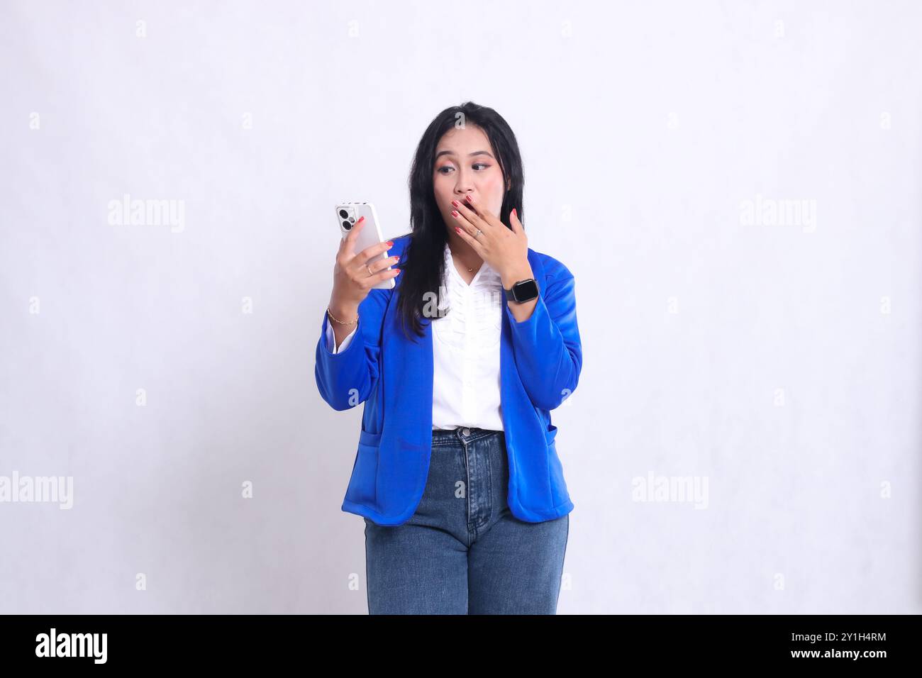 bella ragazza d'ufficio che indossa una tuta formale blu in piedi scioccata che aziona il telefono cellulare dello smartphone mentre la mano si copre la bocca. per tecn Foto Stock