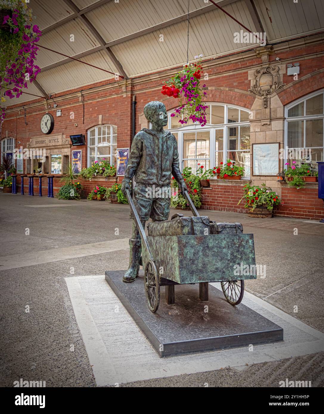 Una statua di bronzo di un barrow boy si trova nel foyer di una stazione ferroviaria. I fiori sono in piantatrici e cesti sospesi e un orologio è sopra l'ufficio prenotazioni Foto Stock