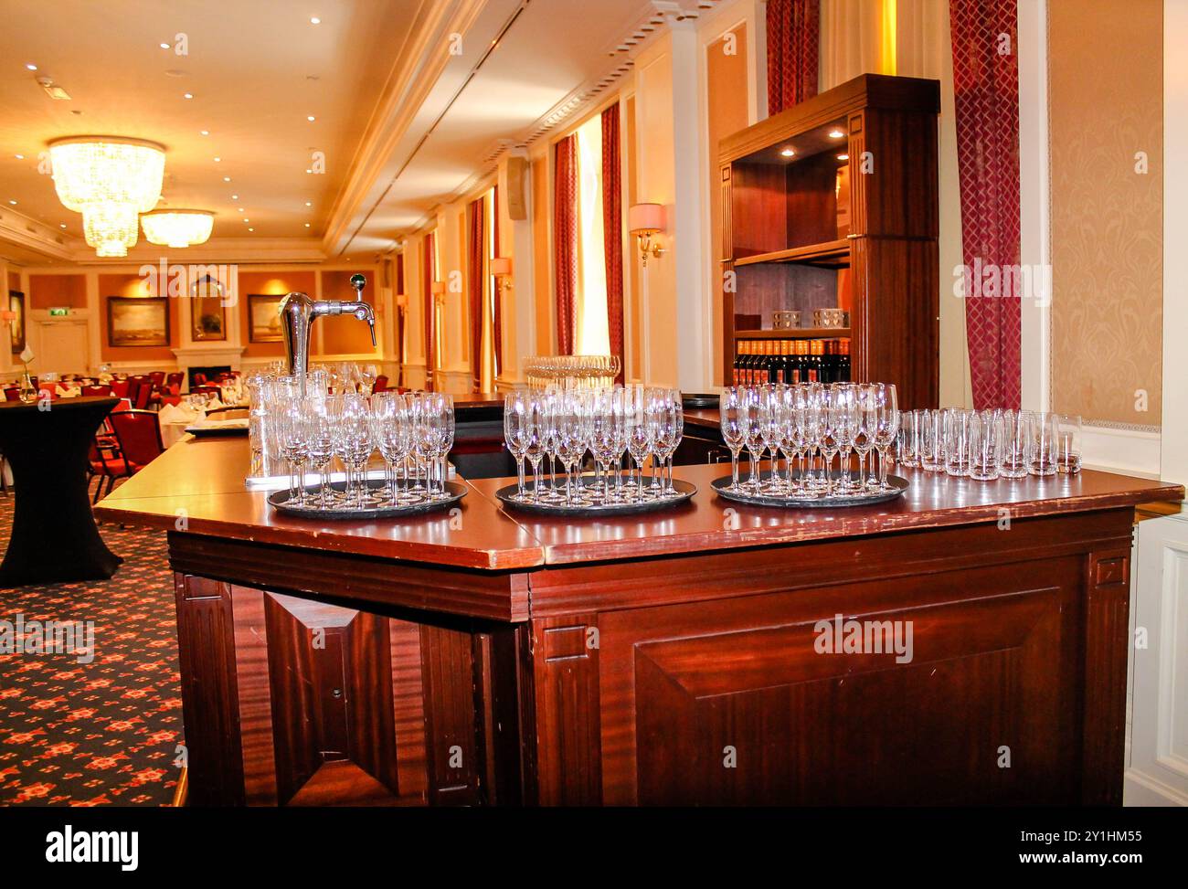 Un'area bar splendidamente arredata con diversi vassoi di bicchieri scintillanti, allestita in un'elegante sala banchetti. La calda illuminazione e il lussuoso decor creano Foto Stock