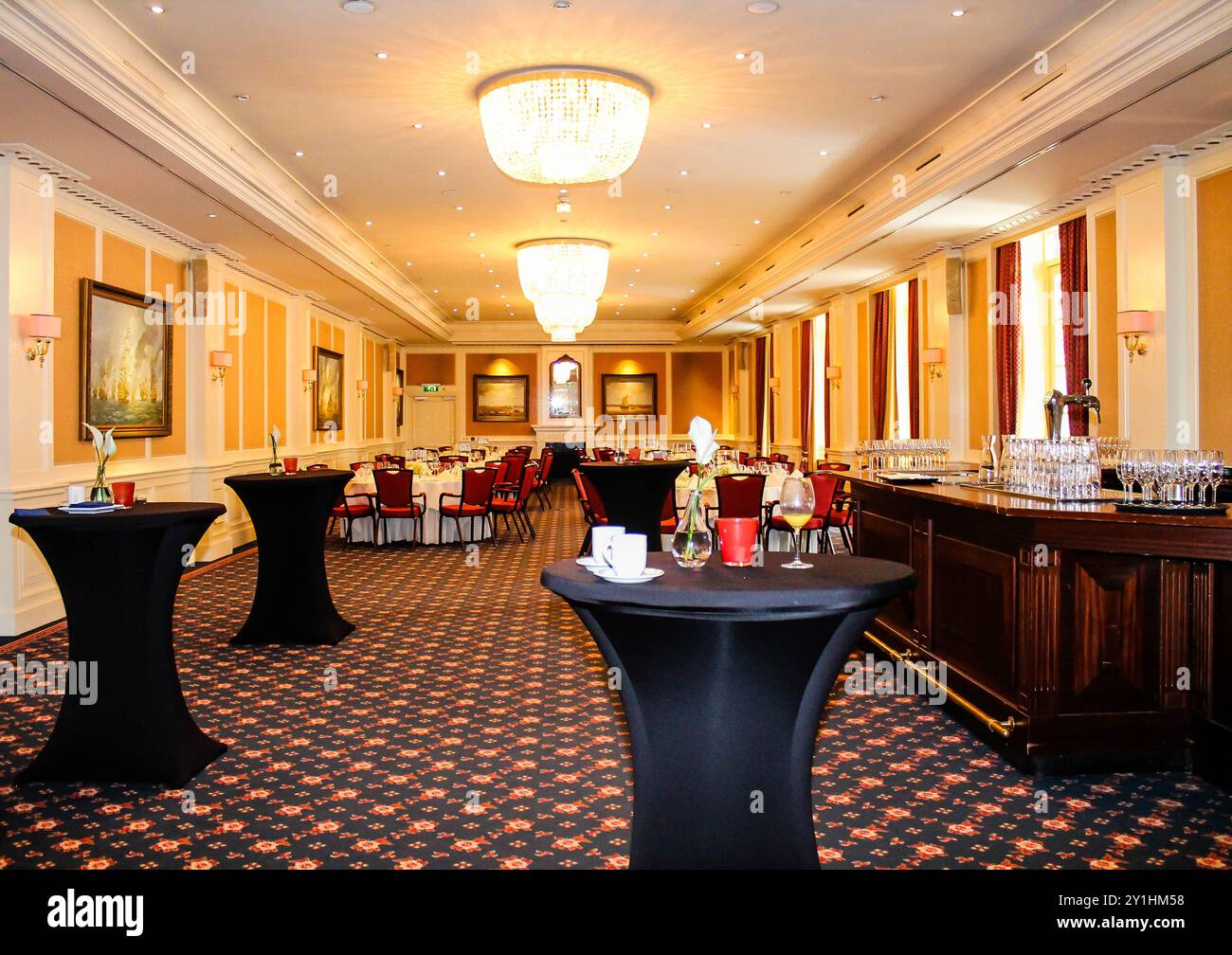 Elegante sala banchetti con tavoli rotondi e tovaglie nere, con area bar e lampadari decorativi. La camera e' ben illuminata con colori caldi a. Foto Stock