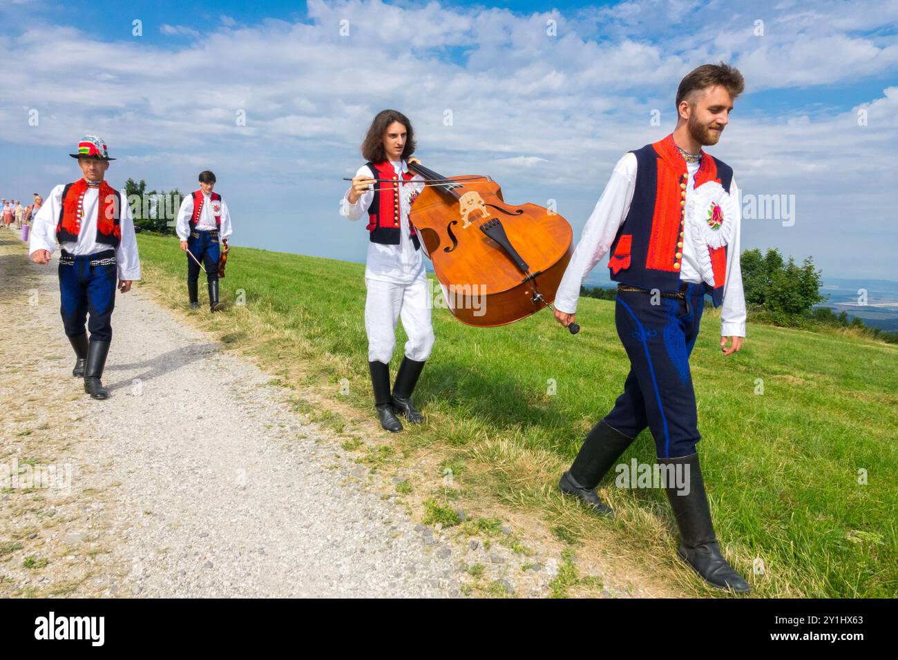 Un gruppo di musicisti in abbigliamento tradizionale cammina su un sentiero di campagna con un contrabbasso sotto un cielo blu Foto Stock