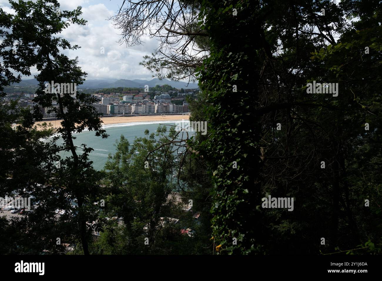 La famosa spiaggia di la Concha con vista dal sentiero che porta al castello, incorniciata dai rami verdi degli alberi. L'acqua è blu, il cielo è nuvoloso, il cit Foto Stock