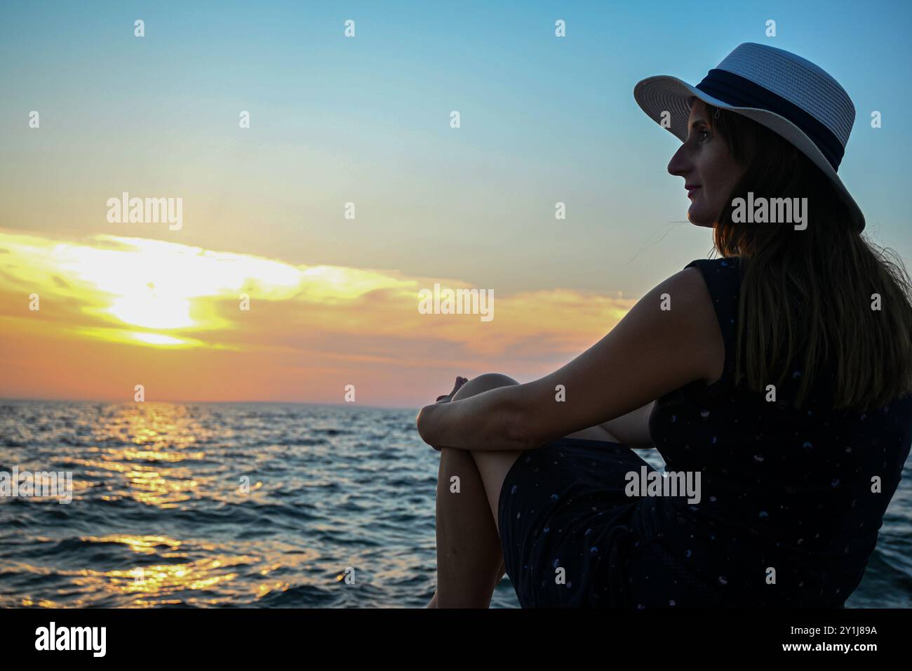 Una bella donna che indossa un abito estivo e un cappello da sole posa al tramonto sulla spiaggia. Foto Stock